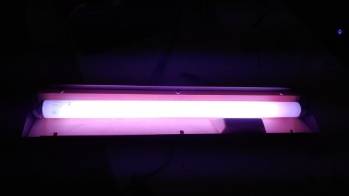 A fluorescent light tube emits a purple glow, placed inside a fixture on a dark surface.