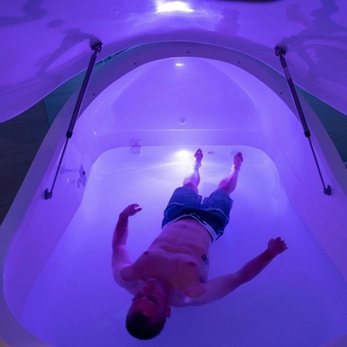 A man floats in a sensory deprivation tank illuminated by soft purple light.