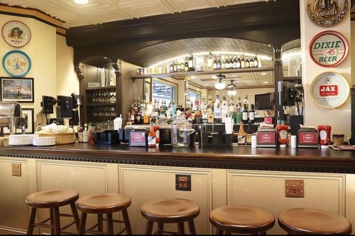 A cozy bar with a wooden counter, stools, and shelves stocked with various bottles and condiments.