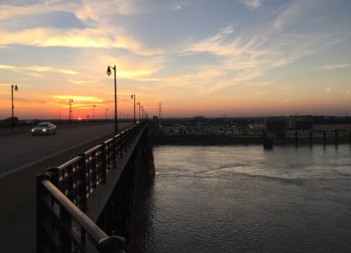A sunset view over a river from a bridge, with a car on the road and clouds in the sky.