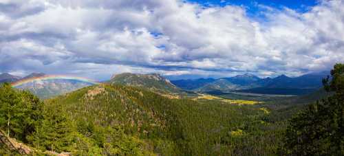 A panoramic view of mountains and valleys under a cloudy sky, featuring a vibrant rainbow arching over the landscape.