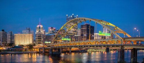 A city skyline at dusk featuring illuminated buildings and a bridge over a river.