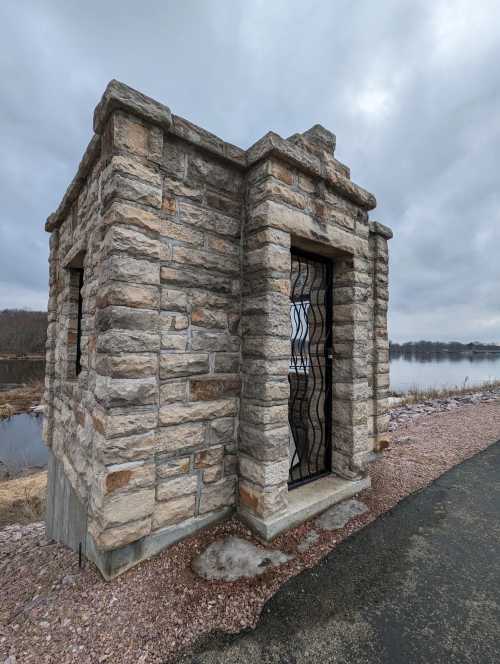 A small stone structure with a metal gate, situated near a body of water under a cloudy sky.