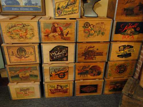 A collection of vintage wooden beer crates featuring colorful labels from Schell Brewing Co. and other brands.