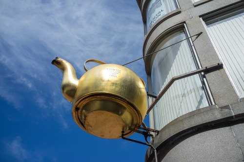 A large golden teapot sculpture mounted on a building, set against a blue sky with scattered clouds.