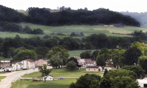 A small town nestled in green hills, featuring houses, trees, and open fields under a cloudy sky.