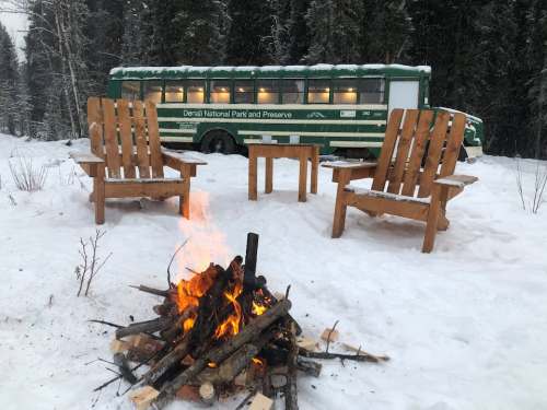 A cozy campfire in the snow with wooden chairs, set against a green bus in Denali National Park.