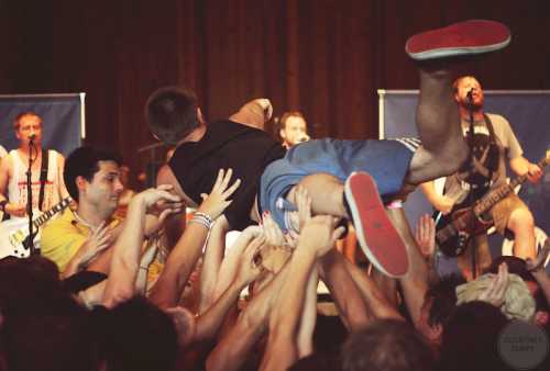 A crowd surges as a concertgoer crowd surfs above eager hands, with a band performing in the background.