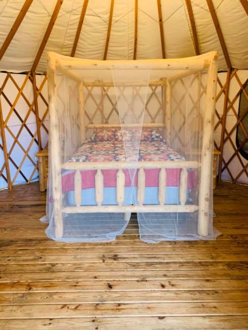 A cozy bed with a mosquito net inside a wooden yurt, featuring a floral-patterned blanket and wooden flooring.