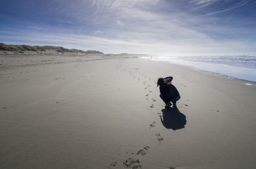 A person crouches on a sandy beach, with footprints leading away and gentle waves in the background under a blue sky.