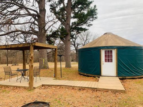 A green yurt with a wooden deck and a small seating area under a pergola, surrounded by trees and grass.