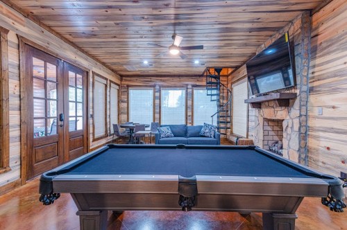 A cozy game room featuring a pool table, wooden walls, a fireplace, and a seating area with a TV.