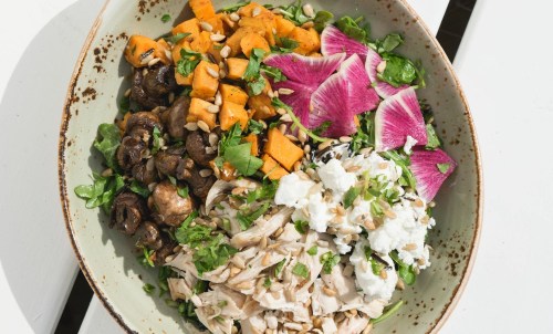 A colorful bowl featuring roasted sweet potatoes, mushrooms, greens, chicken, and pink radish, topped with herbs and seeds.