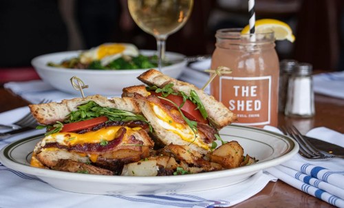 A hearty sandwich with bacon, egg, and greens, served with crispy potatoes and a drink in a cozy restaurant setting.