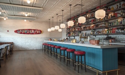 A stylish restaurant interior featuring a bar with red stools, shelves of bottles, and decorative lighting.