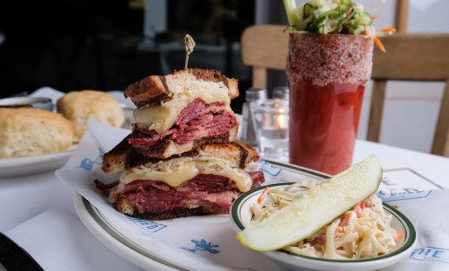 A towering pastrami sandwich with melted cheese on rye, served with coleslaw, a pickle, and a cocktail.