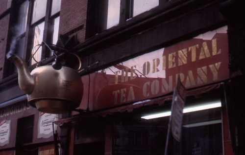 A large brass teapot hangs above the entrance of "The Oriental Tea Company" storefront.