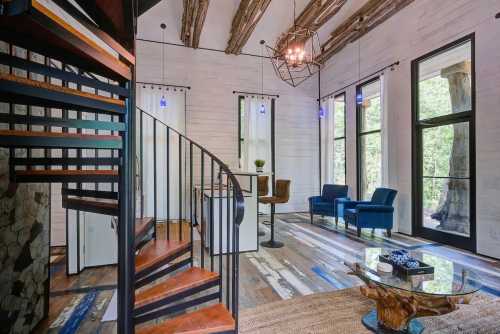 Modern interior featuring a spiral staircase, blue chairs, and large windows with natural light. Rustic wooden beams above.