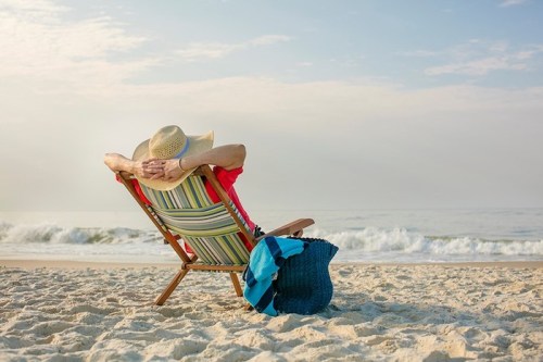 A person relaxes in a beach chair, wearing a straw hat, with the ocean waves gently lapping in the background.