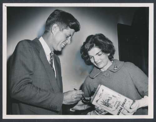 A man in a suit signs a book for a woman in a coat, both smiling in a vintage black-and-white photo.