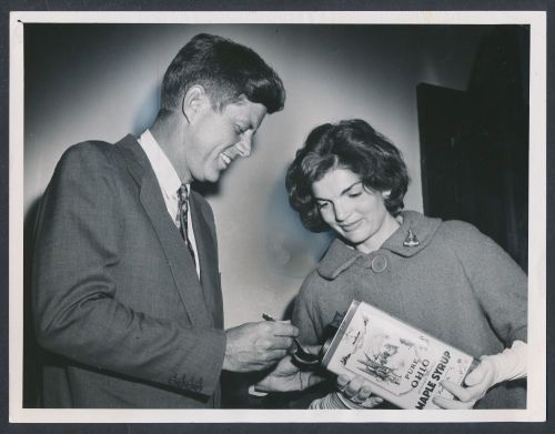 A man in a suit signs a book for a woman in a coat, both smiling in a vintage black-and-white photo.