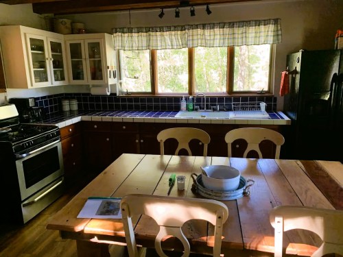 Bright kitchen with wooden dining table, white cabinets, blue tile backsplash, and large window overlooking greenery.