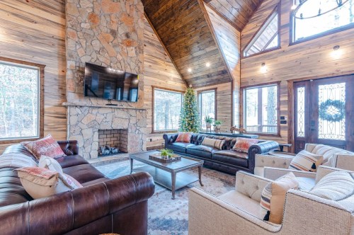 Cozy living room with stone fireplace, wooden walls, and large windows, featuring comfortable sofas and a coffee table.