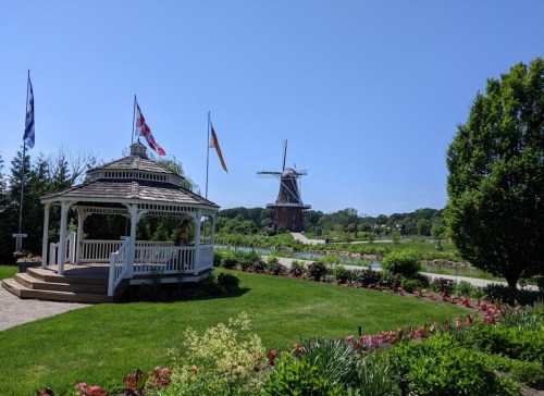 A white gazebo surrounded by green grass and flowers, with a windmill in the background under a clear blue sky.