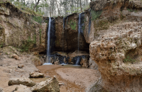 A serene landscape featuring two waterfalls cascading over rocky cliffs into a sandy area below, surrounded by greenery.