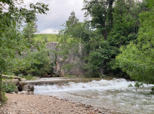 A serene river flows over rocks, surrounded by lush greenery and trees under a cloudy sky.