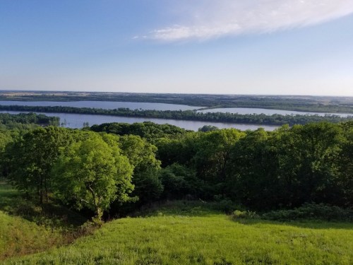 A scenic view of a river surrounded by lush green trees and rolling hills under a clear blue sky.