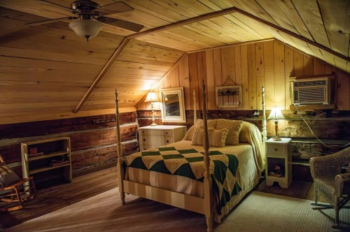 Cozy attic bedroom with wooden walls, a quilted bed, two lamps, and a small sitting area.