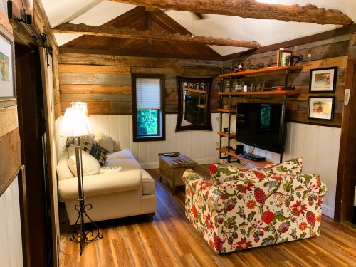 Cozy living room with rustic wooden walls, a white sofa, floral armchair, and a TV on a wooden shelf.