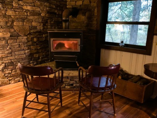 A cozy room with a stone fireplace, two wooden chairs, and a window overlooking trees. Warm and inviting atmosphere.