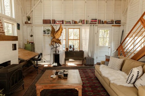 Cozy interior of a rustic cabin featuring a sofa, wooden shelves, and a decorative sculpture. Natural light floods the space.
