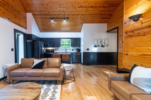 Cozy living area with wooden walls, modern furniture, and a kitchen in the background. Bright and inviting atmosphere.
