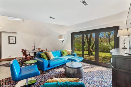 Bright living room with a blue sofa, green cushions, a round table, and large windows overlooking a garden.