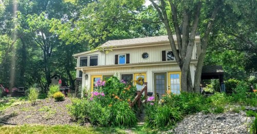 A charming two-story house surrounded by lush greenery and colorful flowers, with a welcoming front porch.