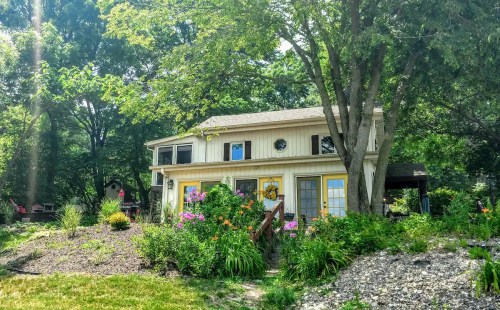 A charming two-story house surrounded by lush greenery and colorful flowers on a sunny day.