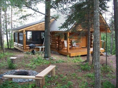 A cozy log cabin surrounded by trees, featuring a porch, outdoor seating, and a fire pit area.