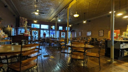 A spacious, empty restaurant with wooden tables, chairs, and vintage decor, featuring large windows and warm lighting.