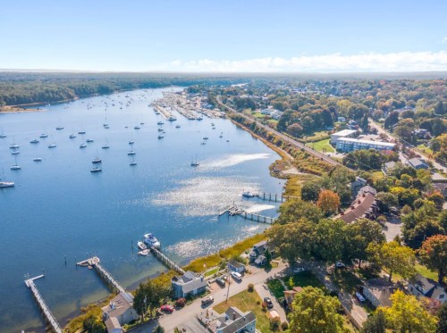 Aerial view of a serene harbor with boats, waterfront homes, and lush greenery under a clear blue sky.