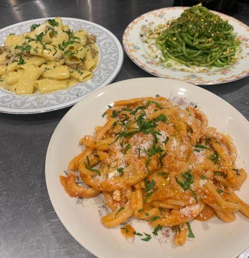 Three plates of pasta: creamy rigatoni, green spaghetti with herbs, and a red sauce pasta topped with cheese and parsley.