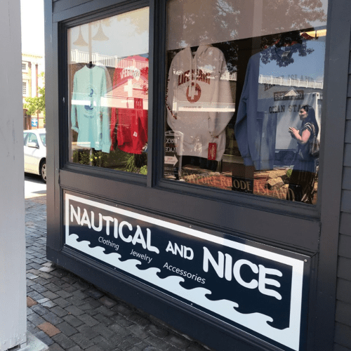 A storefront window displays clothing and accessories with a nautical theme, featuring various apparel and a reflection of a person.