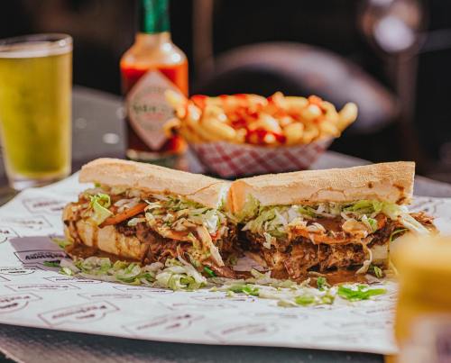 A close-up of a sandwich filled with meat and lettuce, served with fries and a drink in the background.