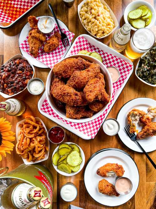 A spread of fried chicken, sides like macaroni and cheese, pickles, and drinks on a wooden table.
