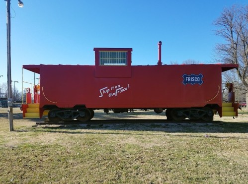 A bright red caboose with "Ship it on the Frisco!" painted on the side, set on grassy ground.