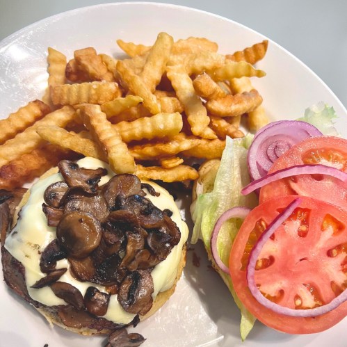 A plate with a burger topped with mushrooms and cheese, served with crinkle-cut fries and a side of lettuce, tomato, and onion.