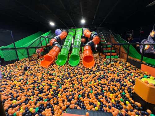 A colorful indoor play area with large slides and a ball pit filled with orange, black, and yellow balls.