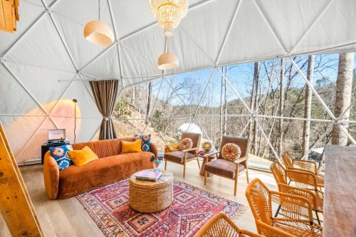 Cozy interior of a geodesic dome with a sofa, chairs, and a view of trees outside. Warm, inviting decor.